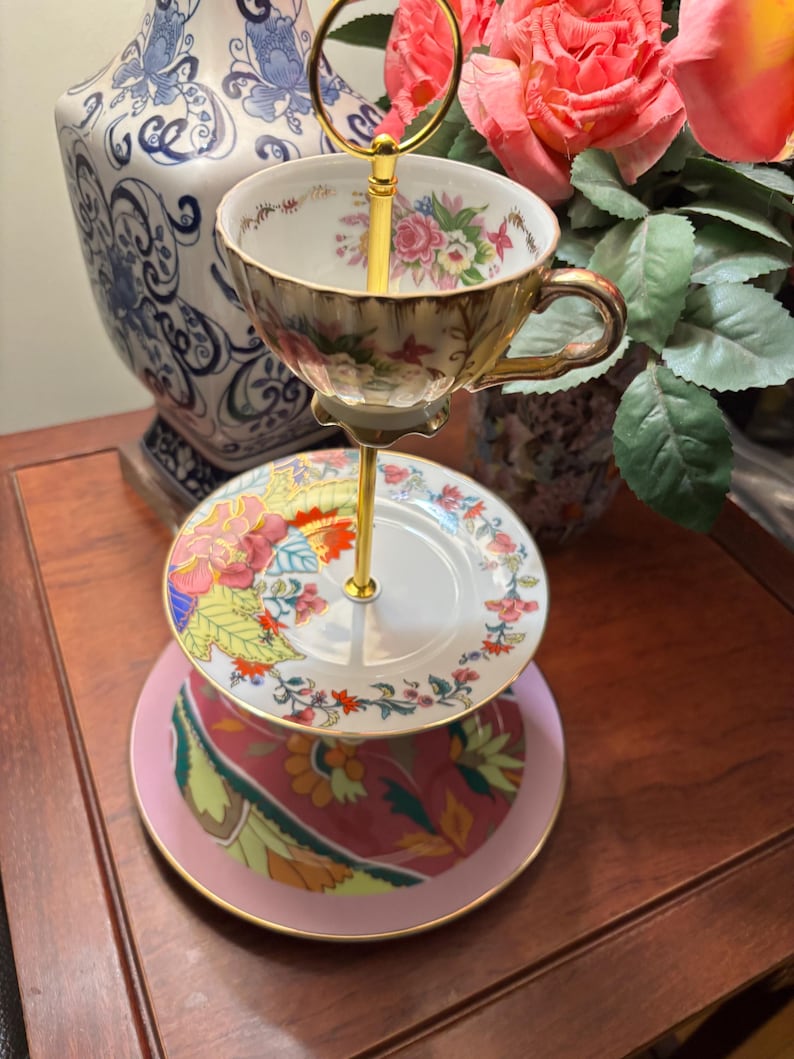 May include: A three-tiered serving stand with a gold handle. The top tier holds a floral teacup. The middle tier is a small plate with a floral design, and the bottom tier is a pink plate with a patterned design. The stand is on a wooden surface.