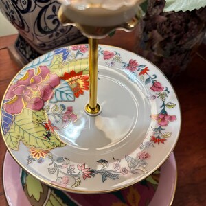 May include: A three-tiered serving stand with floral patterned plates. The plates feature pink, red, and yellow flowers with green leaves. The stand has a gold-coloured central support and a pink base plate.