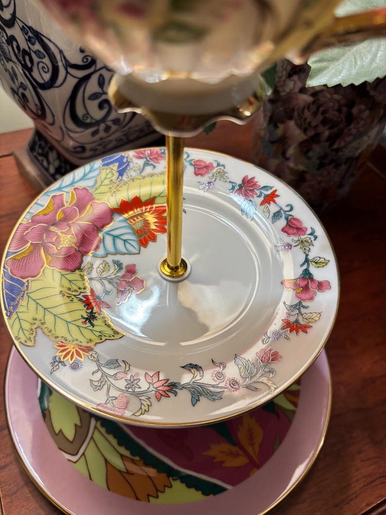 May include: A three-tiered serving stand with floral patterned plates. The plates feature pink, red, and yellow flowers with green leaves. The stand has a gold-coloured central support and a pink base plate.