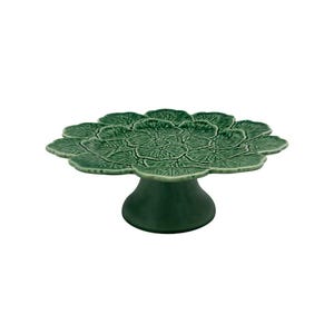 May include: A green ceramic cake stand with a leaf-like design on the top and a pedestal base. The stand is a solid green color and has a textured surface. The cake stand is designed to hold and display cakes or other desserts.