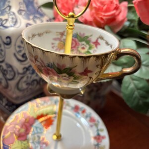 May include: A two-tiered serving stand with a teacup and saucer. The teacup and saucer are white with a floral design and gold trim. The stand is gold-coloured. The background features pink roses and a blue and white vase.