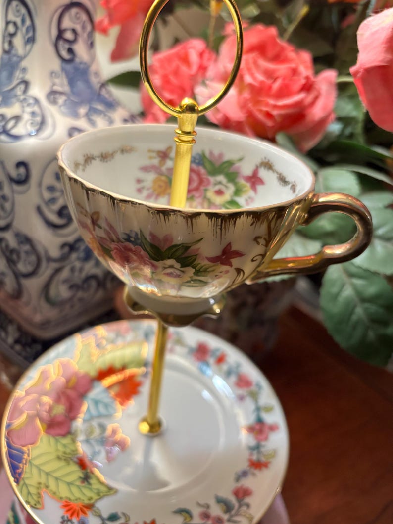 May include: A two-tiered serving stand with a teacup and saucer. The teacup and saucer are white with a floral design and gold trim. The stand is gold-coloured. The background features pink roses and a blue and white vase.