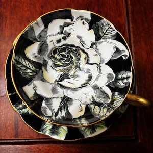 May include: Black and white teacup and saucer set with a large floral design. The teacup and saucer have a gold trim. The floral design is a large white rose with green leaves. The teacup has a gold handle.