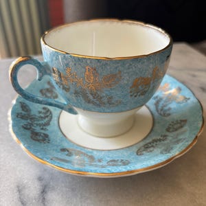 May include: A vintage teacup and saucer set. The cup and saucer are a light blue with gold floral designs and trim. The cup has a white interior and a curved handle. The saucer has a scalloped edge.