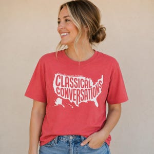 May include: A red short-sleeved t-shirt featuring a white graphic of the United States map with the text "CLASSICAL CONVERSATIONS" printed in large capital letters across the center, along with smaller text above the map.