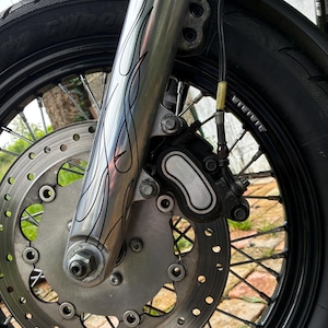 May include: Close-up of a motorcycle front wheel. The wheel has a black rim with silver spokes and a disc brake. The front fork is silver with a flame design. The tire is black.