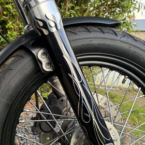 May include: Close-up of a motorcycle front end. The black front fork has a silver flame design. The tire is black and the wheel has silver spokes. The fender is black and the fork is chrome.