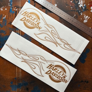 Peut inclure: Deux autocollants Harley-Davidson blancs avec un logo et un motif de flamme marron. Une règle métallique avec des mesures en centimètres est placée en diagonale. Les autocollants sont sur une surface en bois avec des éclaboussures de peinture.