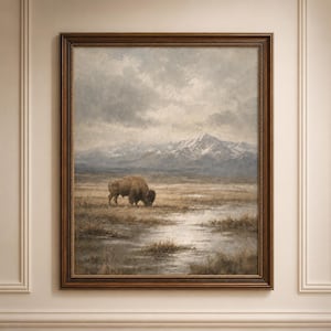 May include: A framed landscape painting depicting a brown bison grazing in a grassy field with a shallow pool of water in the foreground. In the distance, snow-capped mountains rise under a cloudy, overcast sky. The artwork is encased in a dark wood frame.