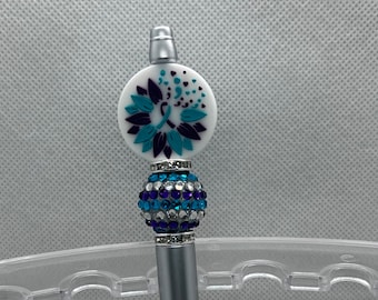May include: A silver pen with a white and purple flower charm with a blue ribbon. The charm is surrounded by clear and blue rhinestones.