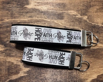 May include: Two black keychains with white ribbon featuring the words "Love, Faith, Hope, Fight" and "Brain Tumor Awareness." The keychains have silver-colored metal hardware and are displayed on a wooden surface.