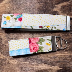 May include: Two fabric keychains with silver key rings. The keychains are made of patchwork fabric with a floral pattern and white polka dots. One keychain has a blue, yellow, and white floral pattern, and the other has a pink and white floral pattern.