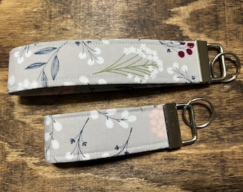 Könnte beinhalten: Zwei graue Stoffschlüsselanhänger mit Blumenmuster und silbernen Schlüsselringen. Die Schlüsselanhänger sind unterschiedlich groß.