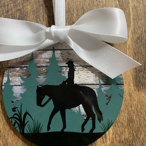 May include: A round, black and green Christmas ornament with a silhouette of a person riding a horse in a forest setting. The ornament is tied with a white ribbon.