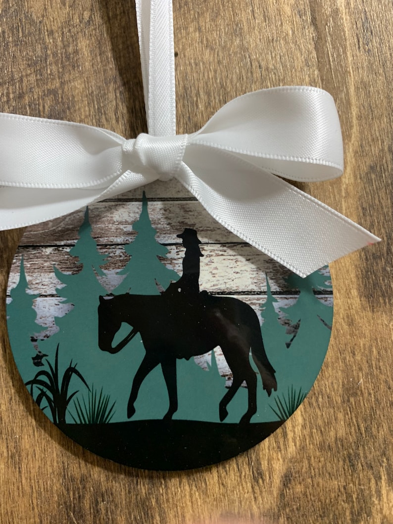 May include: A round, black and green Christmas ornament with a silhouette of a person riding a horse in a forest setting. The ornament is tied with a white ribbon.