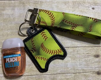May include: A green and brown softball-themed keychain with a small bottle of hand sanitizer. The hand sanitizer bottle is labeled "PocketBac Bath & Body Works Peach Bellini Antibacterial Hand Gel 1 fl oz / 29 ml".