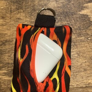 May include: A small black zippered pouch with a flame pattern in orange, yellow, and black. A white wireless earbud case is inside the pouch.