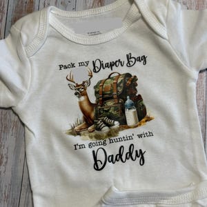 May include: White baby onesie with a deer, backpack, and baby bottle graphic. Text reads "Pack my Diaper Bag, I'm going huntin' with Daddy". The design features a brown deer, a camouflage backpack, and baby shoes.