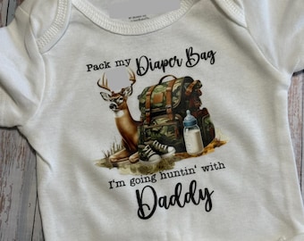 Könnte beinhalten: Weißer Baby-Body mit einem Hirsch-, Rucksack- und Babyflaschen-Motiv. Der Text lautet "Pack my Diaper Bag, I'm going huntin' with Daddy". Das Design zeigt einen braunen Hirsch, einen Tarnrucksack und Babyschuhe.