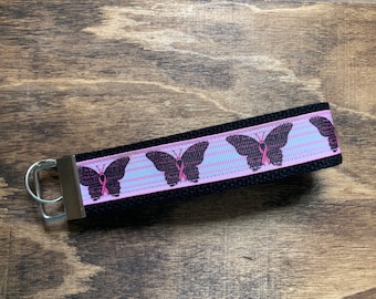 May include: Black and pink key chain with a black butterfly design and pink ribbon accents. The butterfly has a pink ribbon in the center.