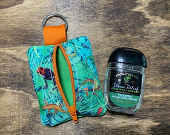 May include: A teal and green zippered pouch with a jungle print featuring frogs and lizards, an orange zipper, and a matching orange loop with a metal ring. Next to it is a 1 fl oz bottle of hand sanitizer.