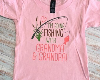Könnte beinhalten: Hellrosa T-Shirt mit dem Text "I'M GOING FISHING WITH GRANDMA & GRANDPA!". Das Design zeigt eine Angelrute, einen Fisch und Schilf. Das Shirt ist aus weichem Material.