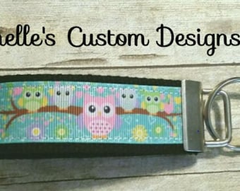 May include: A teal blue and black key chain with a silver key ring. The key chain features a ribbon with a pattern of owls on a branch with pink, green, and yellow flowers. The text "Chelle's Custom Designs" is visible at the top of the image.