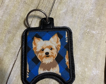 Könnte beinhalten: Ein blauer und schwarzer Stoff-Handdesinfektionsmittelhalter mit einem kleinen Hunde-Bild. Der Halter hat einen schwarzen Lederbesatz und einen silbernen Schlüsselring.