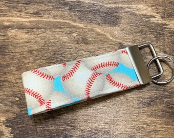 May include: A white fabric keychain with a baseball pattern. The keychain has a silver metal key ring and a silver metal loop.