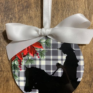 May include: A round Christmas ornament with a black and white plaid pattern. A silhouette of a person riding a horse is in the center of the ornament. The ornament is decorated with red poinsettia flowers and green pine branches.