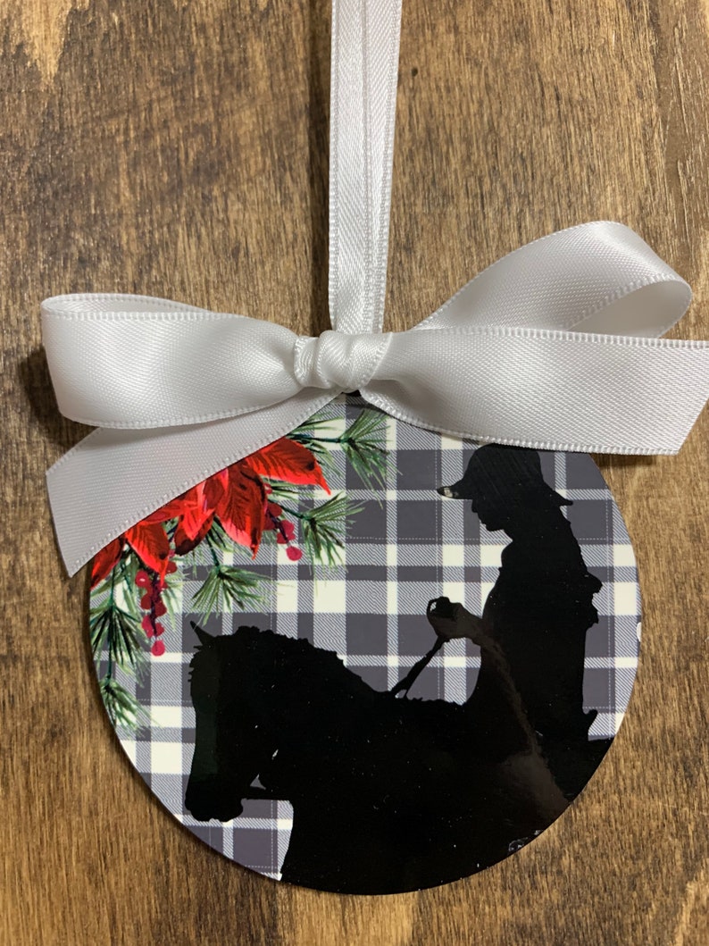 May include: A round Christmas ornament with a black and white plaid pattern. A silhouette of a person riding a horse is in the center of the ornament. The ornament is decorated with red poinsettia flowers and green pine branches.
