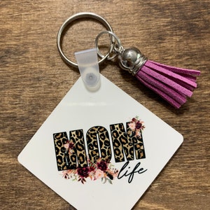 May include: A white square keychain with a pink tassel. The keychain features the word "MOM" in leopard print with floral accents and the text "life" below.