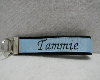 May include: A black and light blue key chain with a silver key ring. The light blue ribbon has the name "Tammie" embroidered in black.