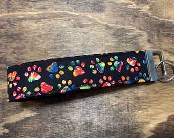 Könnte beinhalten: Ein schwarzer Stoffschlüsselanhänger mit einem Regenbogen-Batik-Pfotenabdruckmuster und einem silbernen Schlüsselring.