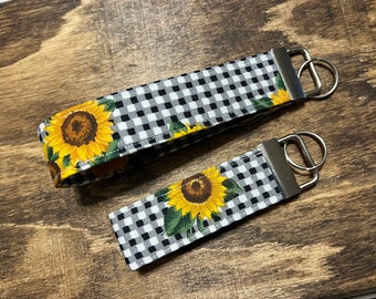 Könnte beinhalten: Zwei Schlüsselanhänger aus schwarzem und weißem Gingham-Stoff mit Sonnenblumenmotiven. Der größere Schlüsselanhänger hat eine einzelne Sonnenblume und der kleinere Schlüsselanhänger hat eine einzelne Sonnenblume.