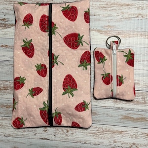 May include: A pink quilted fabric set featuring a large rectangular pouch and a smaller coin purse with a key ring. Both items are decorated with a repeating pattern of red strawberries with green leaves. The pouch has a black zipper.
