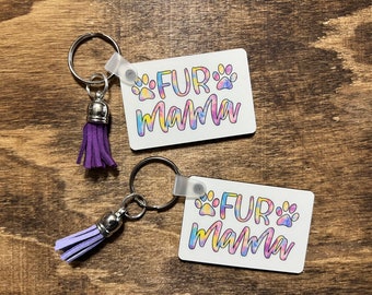 Könnte beinhalten: Zwei Schlüsselanhänger mit violetten Quasten und weißen rechteckigen Anhängern. Die Anhänger haben einen Batik-Hintergrund mit dem Text "FUR mama" und einem Pfotenabdruck auf jeder Seite.