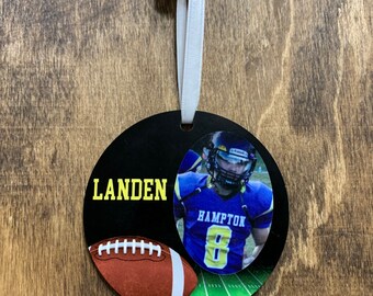 May include: A black and green round ornament with a football player wearing a blue and yellow jersey with the word "HAMPTON" and the number "8" on it. The ornament also has a brown football and the name "LANDEN" in yellow letters.