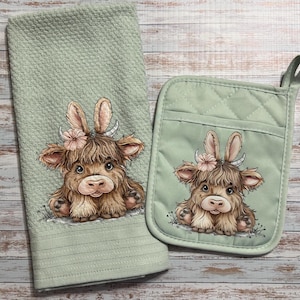 May include: A sage green kitchen towel and oven mitt set. Each item features a whimsical illustration of a Highland cow with bunny ears and a floral crown. The towel has a textured weave, and the mitt is quilted.