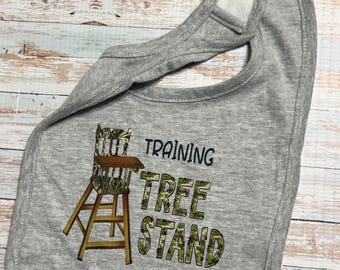 May include: A heather gray baby bib with a hook-and-loop closure at the neck. The bib features a graphic of a wooden high chair with a camouflage pattern and the black text "TRAINING TREE STAND" with the words "TREE STAND" in camouflage.