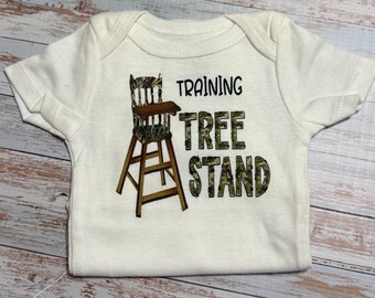 Könnte beinhalten: Ein weißer kurzärmeliger Babybody mit einer Grafik eines Hochstuhls im Camouflage-Muster. Der Text "TRAINING TREE STAND" ist in schwarzen und tarnfarbenen Blockbuchstaben neben der Abbildung des Hochstuhls aufgedruckt.