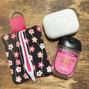 May include: A black floral print zippered pouch with a pink zipper and a pink strap, a white AirPod case, and a pink Bath & Body Works hand sanitizer bottle with the text "A Thousand Wishes".