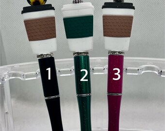 May include: Three decorative pens with coffee cup toppers. The pens have black, green, and magenta barrels, each with a silver tip. The toppers feature a sunflower, green, and red bead design. The pens are displayed in a clear plastic holder.