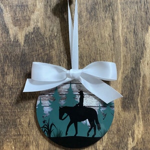 May include: A round, white, wooden ornament with a green and white forest scene and a silhouette of a person riding a horse. The ornament has a white ribbon tied around it.