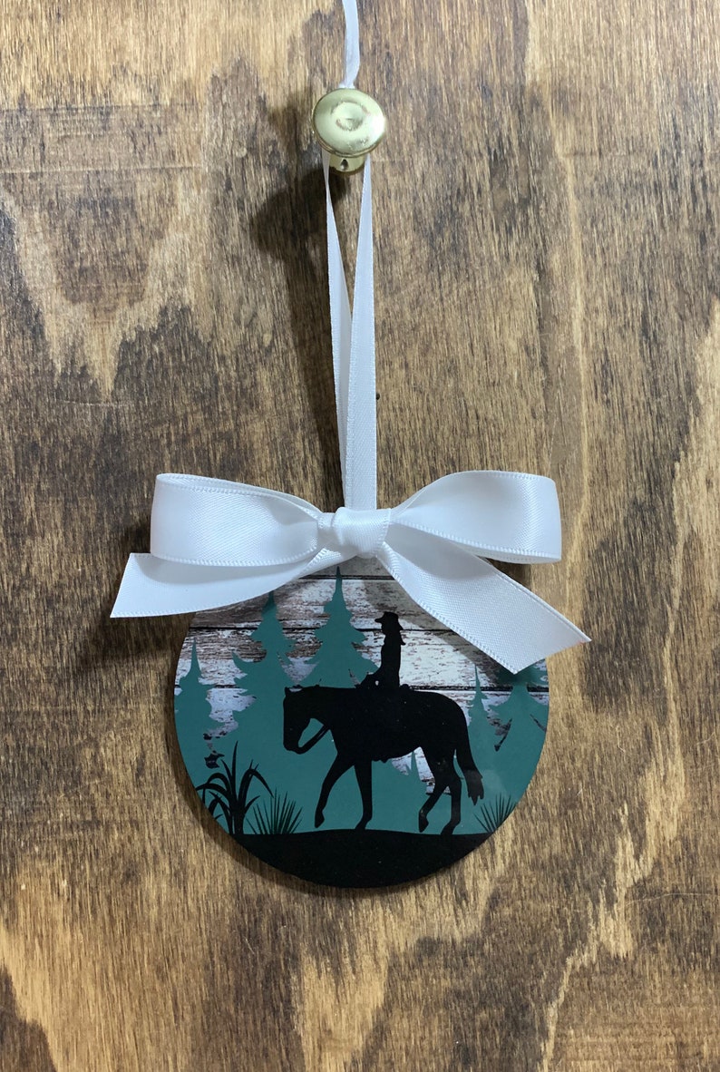 May include: A round, white, wooden ornament with a green and white forest scene and a silhouette of a person riding a horse. The ornament has a white ribbon tied around it.