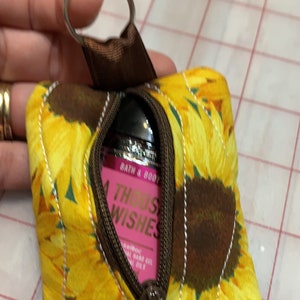 May include: A small zippered pouch with a sunflower print. The pouch is brown with yellow sunflowers and has a zipper closure. A small bottle of hand sanitizer is inside the pouch. The bottle is pink and says "Bath & Body Works" and "A Thousand Wishes".