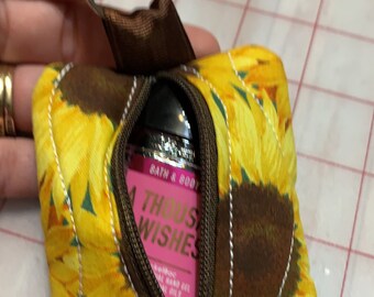 May include: A small zippered pouch with a sunflower print. The pouch is brown with yellow sunflowers and has a zipper closure. A small bottle of hand sanitizer is inside the pouch. The bottle is pink and says "Bath & Body Works" and "A Thousand Wishes".
