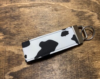 May include: A fabric keychain with a cow print design. The keychain features a white background with black spots, a silver-colored metal clasp, and a key ring. The fabric is rectangular and appears to be durable.
