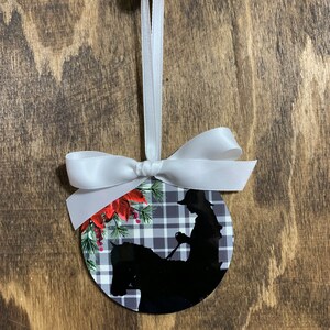 May include: A round Christmas ornament with a black and white plaid background. A silhouette of a person riding a horse is in the center of the ornament. The ornament has red poinsettia flowers and green pine branches around the edge.