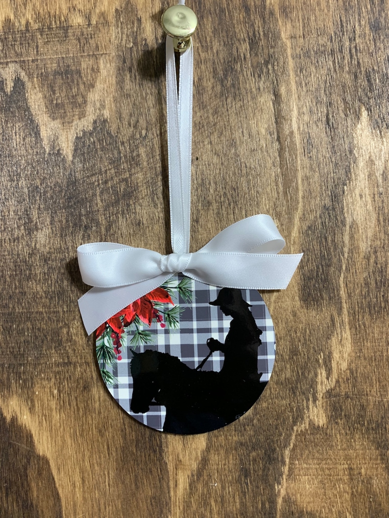 May include: A round Christmas ornament with a black and white plaid background. A silhouette of a person riding a horse is in the center of the ornament. The ornament has red poinsettia flowers and green pine branches around the edge.